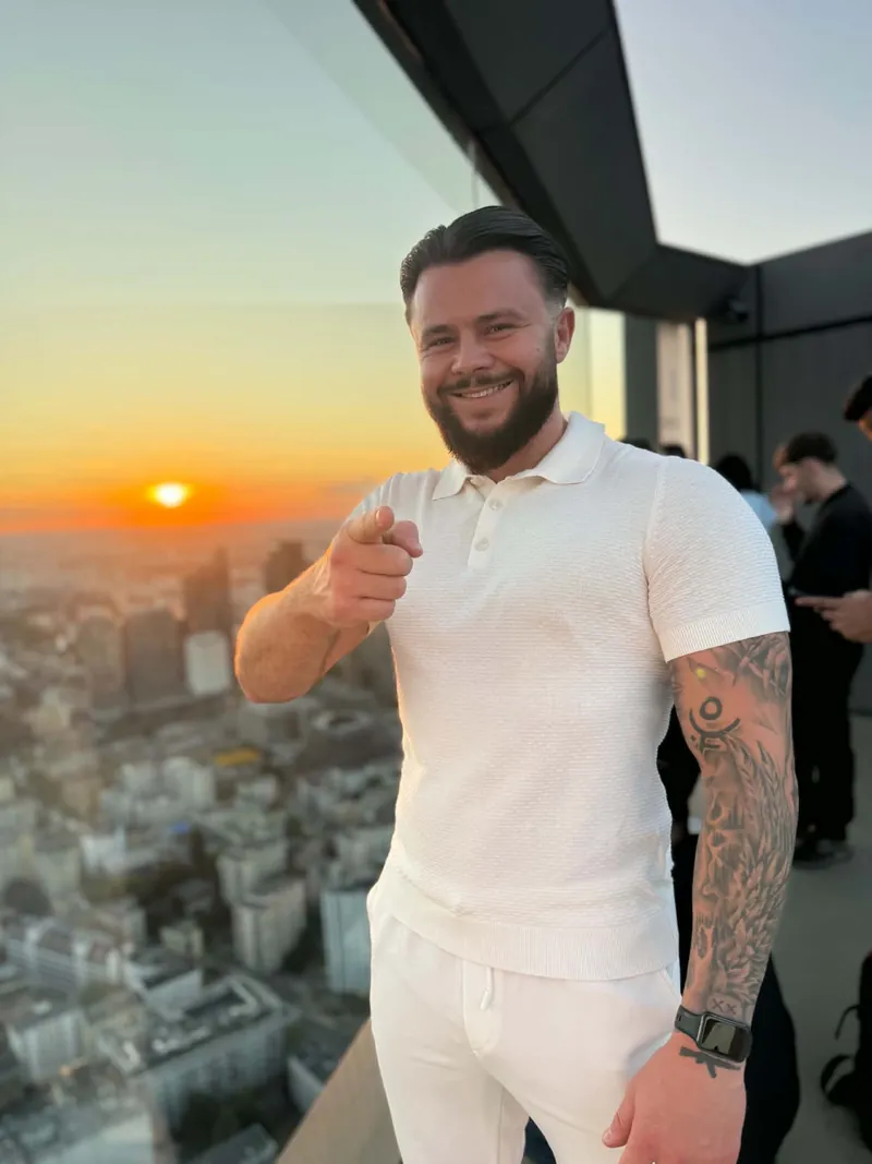 Daniel Grzesiuk w białej koszulce polo uśmiecha się i wskazuje palcem w stronę aparatu